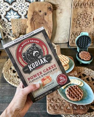 Hand hält Kodiak Power Cakes Buttermilk-Mix mit Proteinwaffeln, Bananen und Waffeleisen auf rustikalem Tisch im Hintergrund