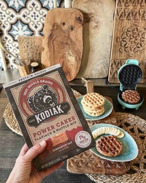 Hand hält Kodiak Power Cakes Zimt-Hafer-Mix mit Proteinwaffeln, Bananen und Waffeleisen auf rustikalem Holztisch im Hintergrund.