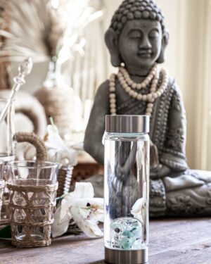 VitaJuwel ViA Edelstein Wasserflasche mit Aquamarin, Aventurin, Rauchquarz und Bergkristall auf einem Boho-Style Holztisch vor einer Buddha-Statue