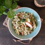 Makrofreundlicher Brokkolisalat mit Apfel, Kichererbsen und Räuchertofu in einer Schüssel auf einem Holzbrett, daneben Basilikum