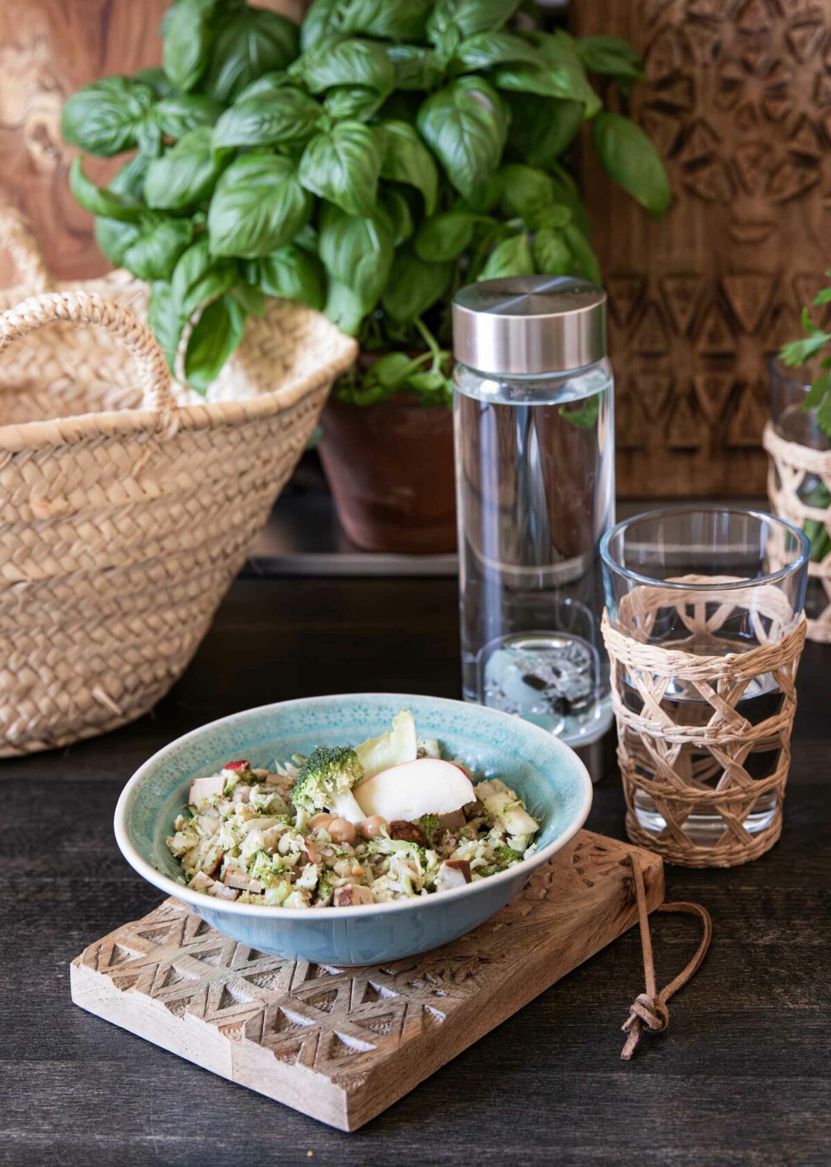 Makrofreundlicher Brokkolisalat mit Apfel, Kichererbsen und Räuchertofu auf einem dekorativen Holzbrett, mit Wasser und Basilikum im Hintergrund