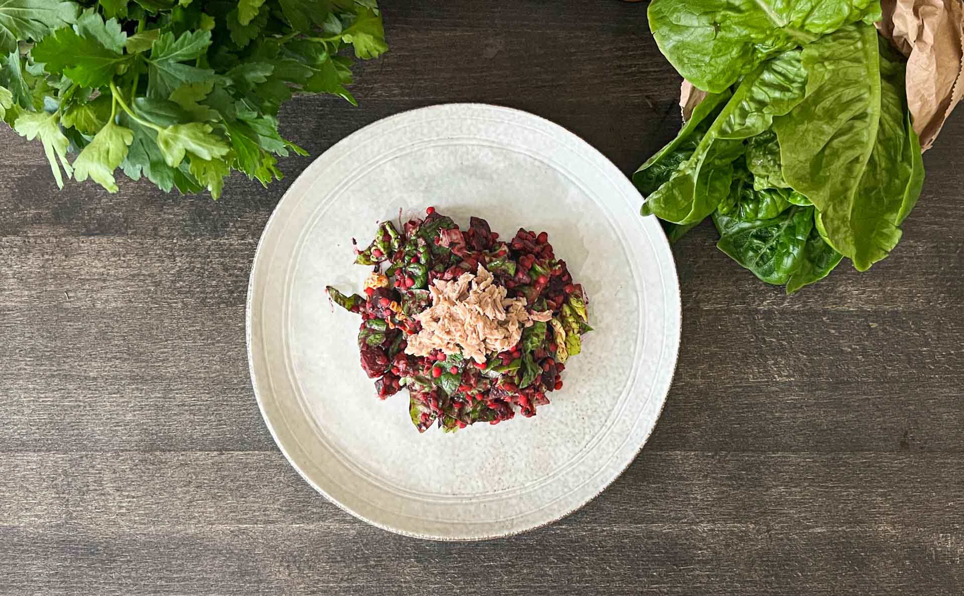 Rote-Bete-Linsen-Salat mit Thunfisch auf einem weißen Teller, umgeben von frischen Kräutern.