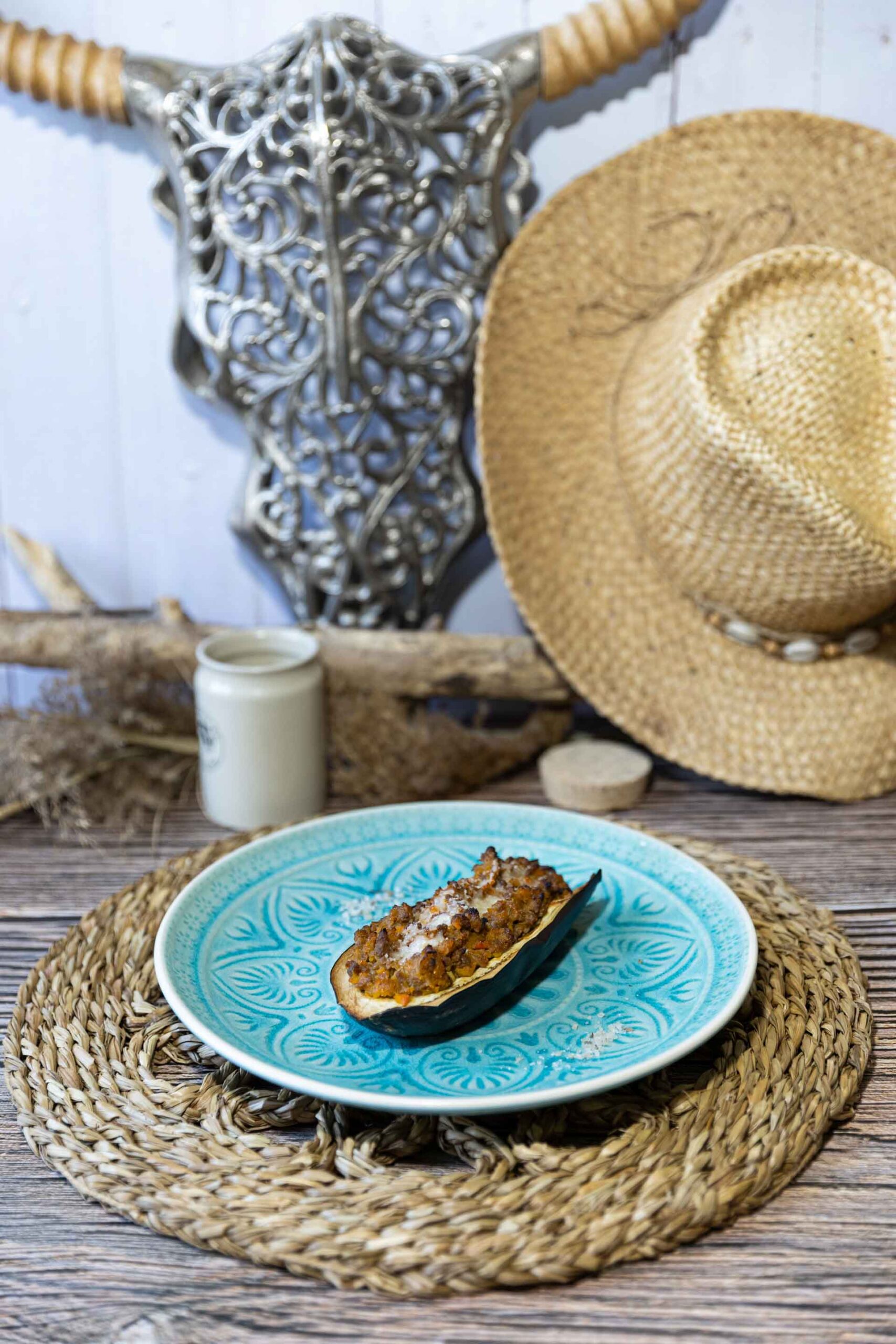 Gefüllte Aubergine mit Hackfleisch auf einem blauen Teller, rustikal dekoriert mit Strohhut und Holzelementen.