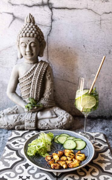 Low-Carb Airfryer Curry-Fischchips mit Gurken und Salat vor einer Buddha-Statue.